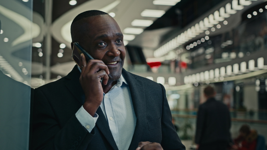 Smiling African American middle-aged man talking mobile phone in company shopping mall. Cheerful adult mature businessman enjoy cellphone call happy entrepreneur calling smartphone indoors speak laugh | Shutterstock HD Video #1097360741