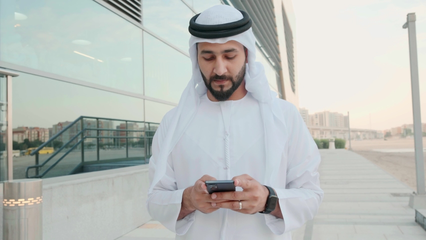 UAE Emirati man using mobile cell phone. Arab businessman wearing cultural Kandura and Ghutra texting, browsing or checking online banking concept. 4K 50p Portrait footage of Arabic guy. | Shutterstock HD Video #1097369549