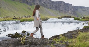 Iceland travel travel video. Woman walking by waterfall on Iceland. Girl tourist in casual clothing visiting icelandic nature landscape. SLOW MOTION