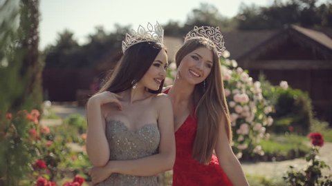 Two sexy young female in long gowns and crowns smiling and posing in the park with roses