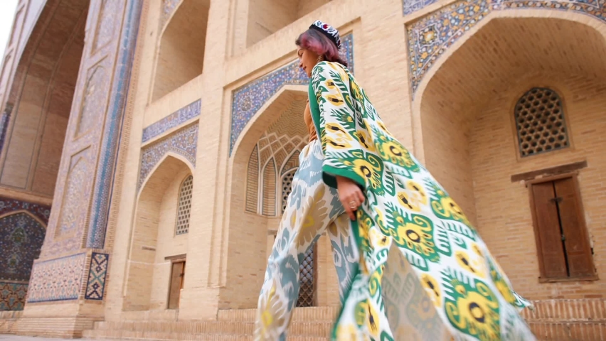An oriental girl in a traditional Uzbek dress walks past an old mosque. Landmark of Bukhara. Back view. Eastern madrasah.