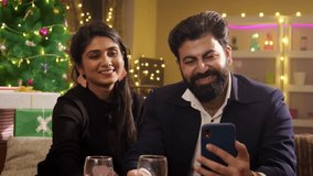 A happy smiling well dressed Indian Asian couple or husband and wife sitting together toasting wine glasses with family or friends talking on a video call using a mobile phone on Christmas festival