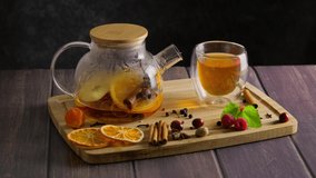 Making hot fruit winter tea with orange and raspberry at home, woman's hand putting a cinnamon stick into glass with hot drink, glass teapot placed on wooden table, slow motion hd video, 4k footage