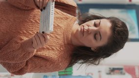 Vertical video: Woman examining drugstore shelves with pharmaceutical products, looking for prescription treatment to buy medicaments. Female client checking pills and boxes at pharmacy. Handheld shot