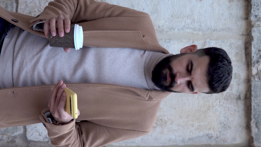 Bearded young man in a jacket and turtleneck nodding his head in agreement at something he has seen on his mobile phone while taking a sip of his portable coffee.