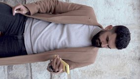 Vertical video of a young bearded man in a jacket and turtleneck putting on a wireless earphone while manipulating music with his cell phone and moving his head.