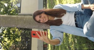 Vertival video.Smiling Spanish young woman making a video call in a park with a smart phone
