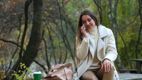 Caucasian woman sits on the wooden table in the park her bag beside her. Calm woman speaks on the phone in autumn park. Blurred backdrop.