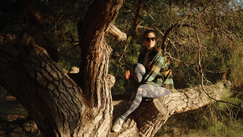 A girl with a scythe in a plaid shirt and glasses, sitting on a pine tree in the forest and looking at the sun.