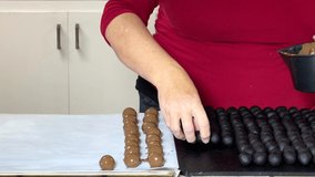 4K HD video of an older caucasian female in kitchen dipping cake balls in chocolate candy melted, then dropping coated cake balls with a fork on a cookie sheet to set up.

