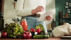 Young woman standing in kitchen and checking writing a shopping list on a notebook at home. Healthy Food on Table, Vegetable Salad recipe, Diet. Dieting Concept, Cooking At Home. - Powered by Shutterstock - Get 15% off with code: PIKWIZARD15