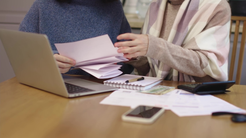 Two female in warm clothes sitting kitchen table look through papers bills documents counting calculator expenses payments utility fees taxes. laptop smartphone on table. concept home budget problems | Shutterstock HD Video #1097400511