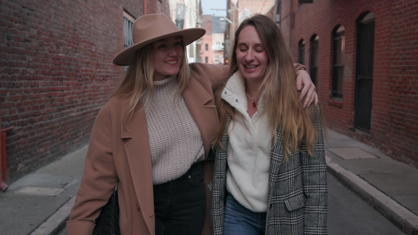 Close-up of cheerful, young women in stylish, autumn outfits. Best girlfriends strolling along narrow city lane and laughing while gossiping. High quality 4k footage