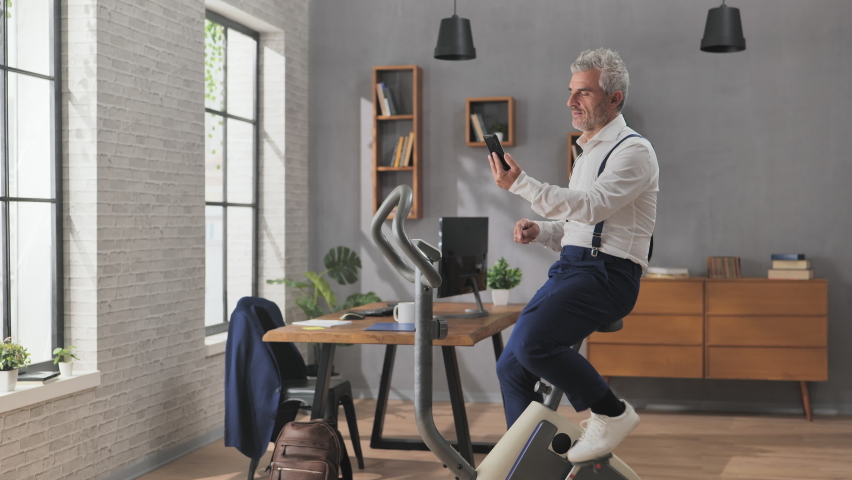 Businessman has a work video conference call in home office while he does exercise gym activity on stationary bycicle,multitasking business man | Shutterstock HD Video #1097402509
