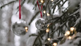 Two silver faceted balls hang on natural branches of snow-covered spruce decorated with warm light garland and spin, close-up. Christmas toys hang on red ribbon without people, view up. - Powered by Shutterstock - Get 15% off with code: PIKWIZARD15