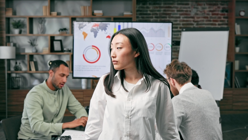 Confident happy smiling diverse asian woman, startup team leader, standing against background of mixed race office workers or business people with arms crossed or clasped, looking at camera. | Shutterstock HD Video #1097403557