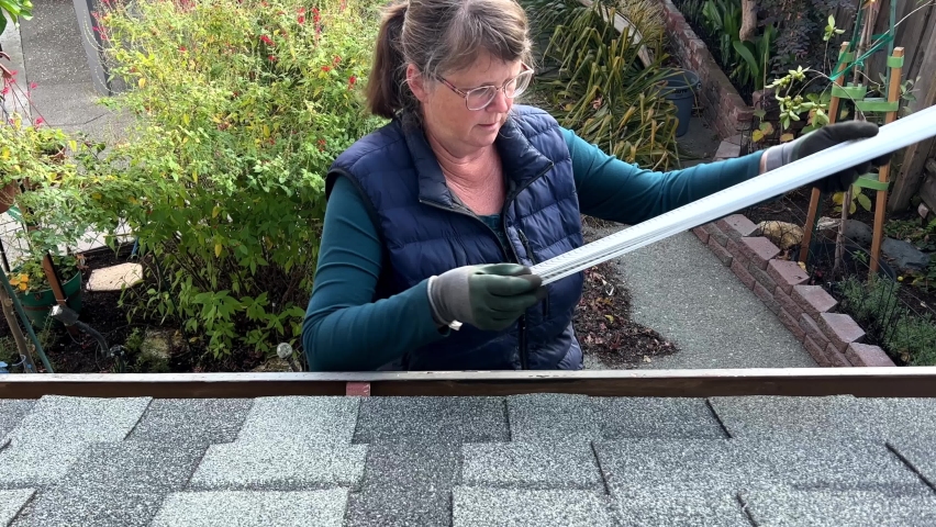 4K HD video of an older caucasian woman wearing basic gardening gloves installing gutter guards under shingles of roof over rain gutters on house. view from above with backyard in background.
 Royalty-Free Stock Footage #1097404535