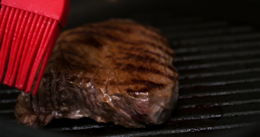 Extreme close up shot of a delicious fried steak in the hot pan being brushed with oil with a red brush for a delicious dinner. Slow Motion