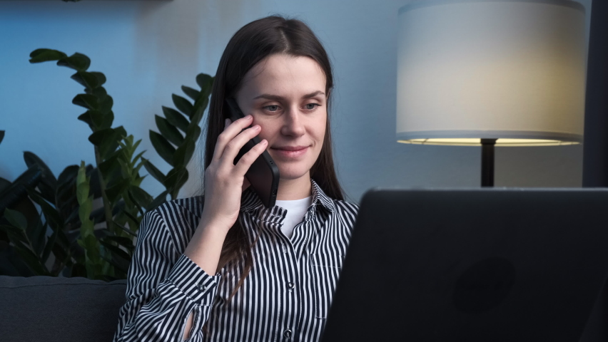 Happy beautiful female customer make mobile call confirming online website shopping order delivery concept. Smiling young businesswoman professional talking on phone using laptop sit on sofa ta home | Shutterstock HD Video #1097414409