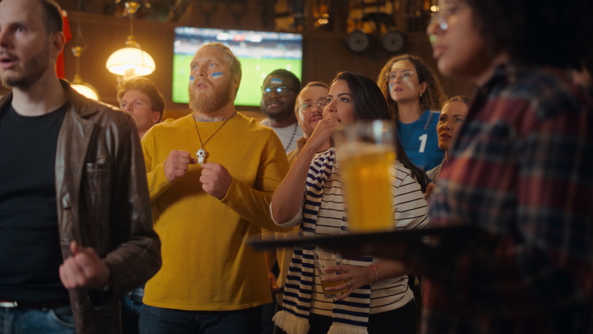 Group of Multiethnic Soccer Fans Watching a Live Football Match in a Sports Bar. People Standing in Front of a TV, Nervous for Their Team. Crowd Disappointed When Opponents Score a Goal. Slow Motion.