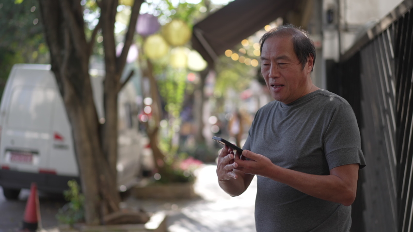 One Senior Asian man talking on phone standing outdoors in street sidewalk. Casual older person speaking on smartphone in city