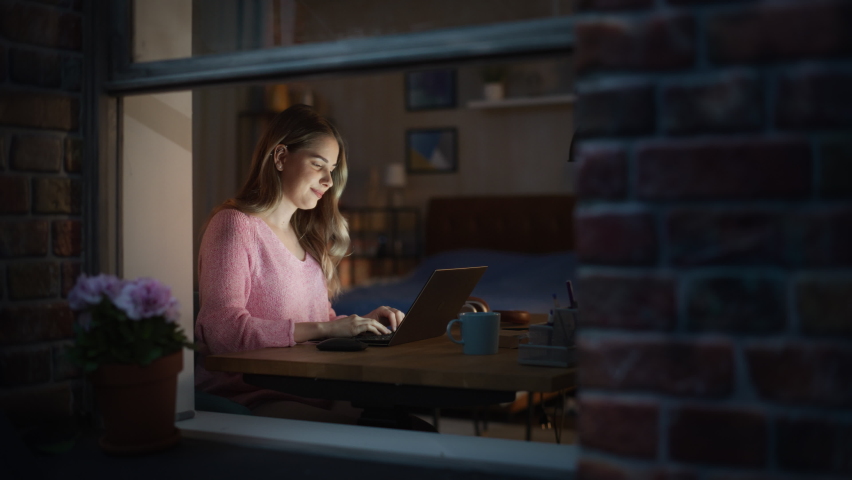 Portrait Shot of a Beautiful Smiling Young Woman Working on a Laptop in a Cozy Bedroom. Smiling Caucasian Female Using Searching Engines and Browsing the Internet. View Through the Window From Outside