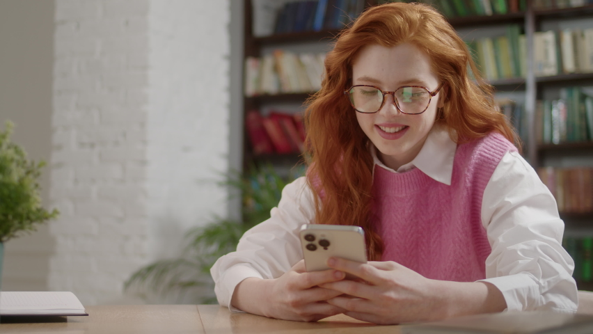 Portrait of happy young student chatting by mobile phone in university library. Attractive Woman with Red Hair and Eyeglasses using social media application browsing on smartphone. Library Background Royalty-Free Stock Footage #1097426045