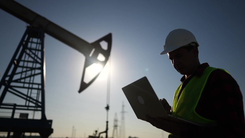 Oil industry. a worker works next to an oil pump holding a laptop. industry business oil and gas concept. engineer studying the level of oil production on a laptop silhouette at lifestyle sunset