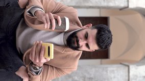 Vertical video of a young bearded man in a jacket and turtleneck sitting while looking at his mobile phone with one hand and drinking coffee with the other.