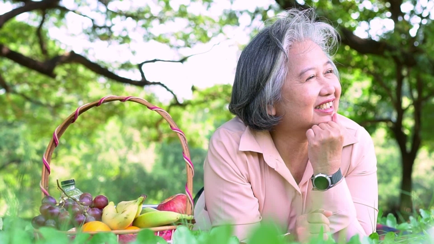 Happy Asian old senior woman and wear a health watch and lying on the picnic mat in park and basket of fruit besides. Concept of happy elderly woman after retirement and good health | Shutterstock HD Video #1097430717