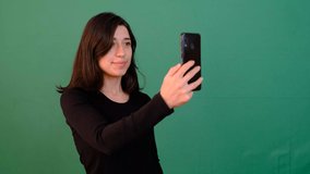 Woman taking selfie on green screen, pretty young girl smiling look smartphone front camera, she is black blouse and short hair in front of chroma key