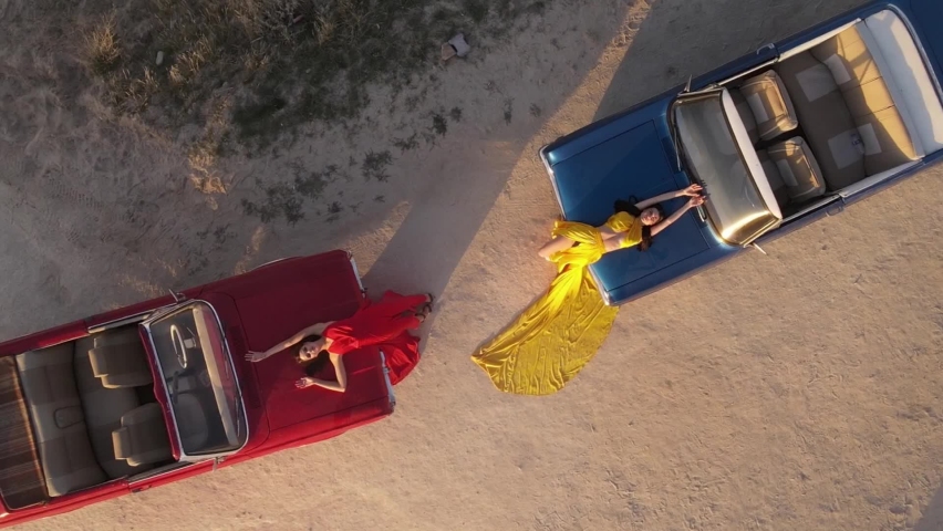 Aerial footage of Two Girls in Dress Lying On Hood Of Cars