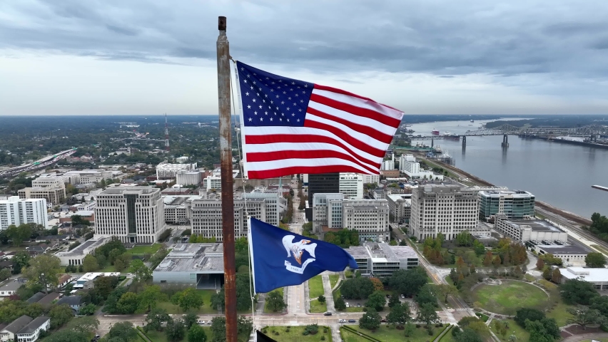USA and Louisiana state flag wave in breeze. Aerial from atop state capitol building. View of downtown Baton Rouge and Mississippi River.