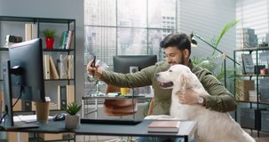 Young handsome Hindu guy is sitting in office and taking selfie with his big dog retriever for social media.