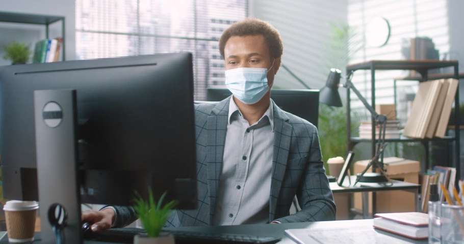 Portrait of successful african american office worker in medical mask sitting in office and taking off protection smiling at camera. Royalty-Free Stock Footage #1097450959