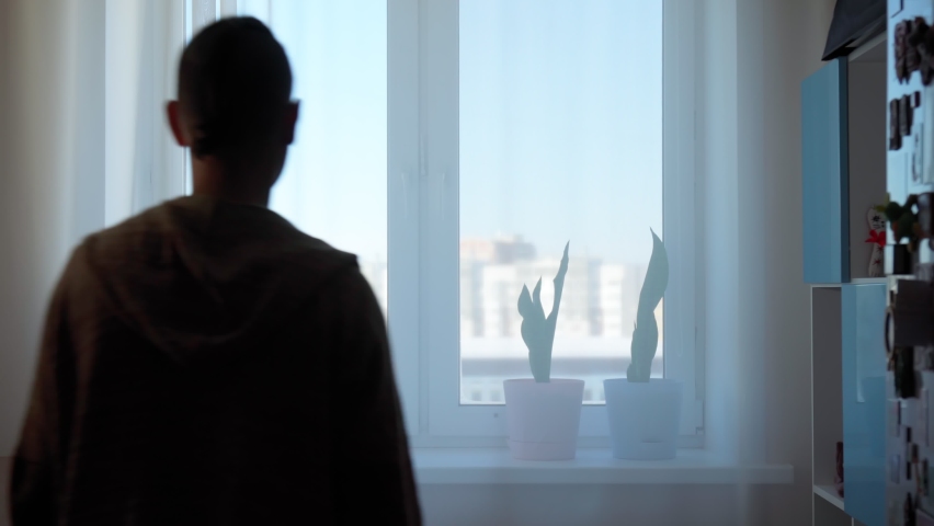 Rear view of a man standing in front of a window and looking out at home. Selective focus