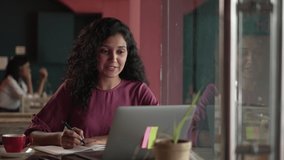Smiling modern Indian freelancer business woman talking online video call laptop working remotely at coffee shop.Asian female looking at the laptop speak explain and writing notes in a notebook.
