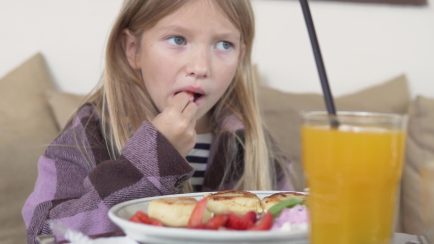 A little girl has breakfast in a restaurant, cottage cheese with strawberries and freshly squeezed orange juice. | Shutterstock HD Video #1097465485