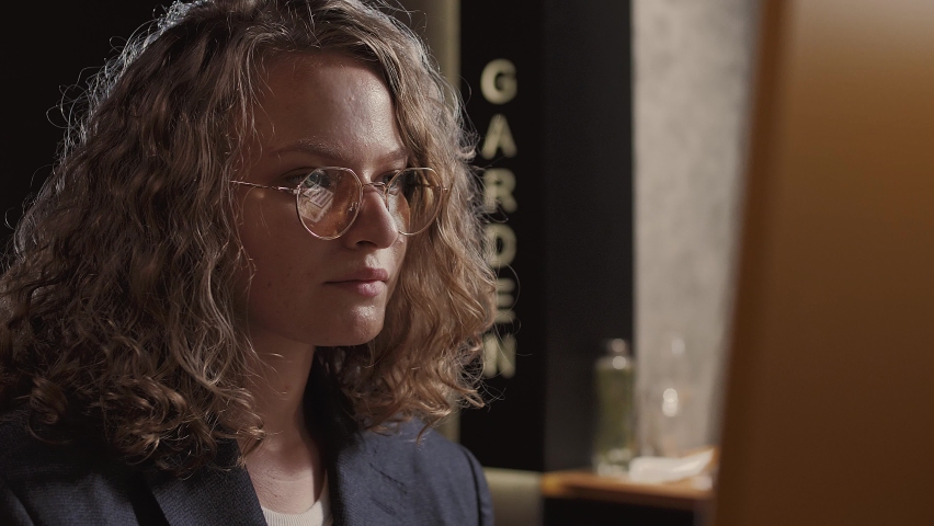 Cute girl graphic designer with glasses and wavy hair sits at her computer and looks at the monitor. Portrait