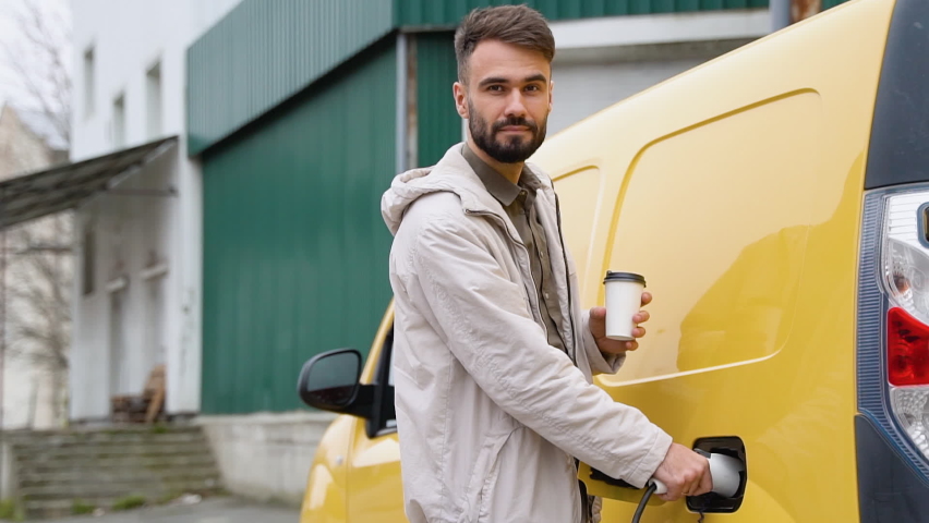 Deliveryman with coffee cup charging cargo electric car near warehouse