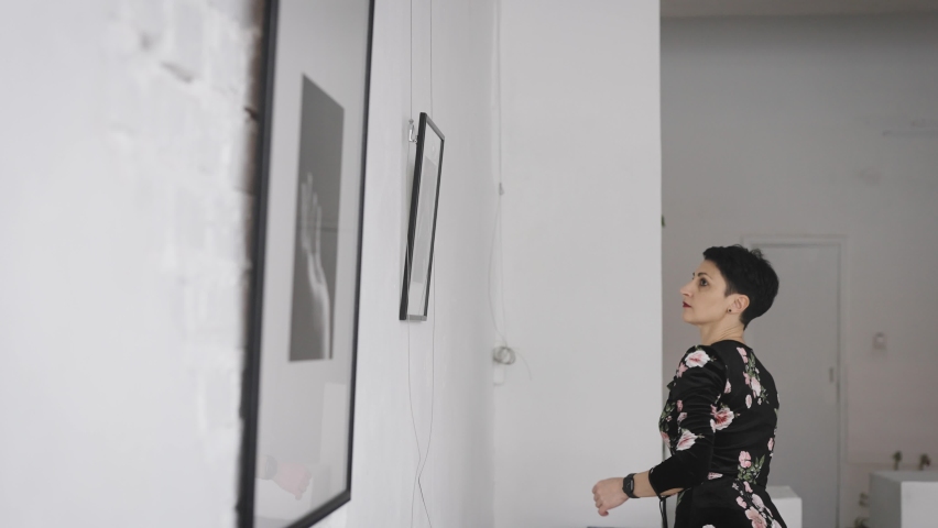 A woman gallerist hangs new paintings and collages on the wall of the exhibition hall. An expert on contemporary art works in galleries and prepares new exhibitions and auctions.