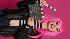 Vertical video: Cheerful female videomaker holding film slate and saying action, preparing to record video montage or footage. Young beautiful person with filming equipment, filmmaker over background.