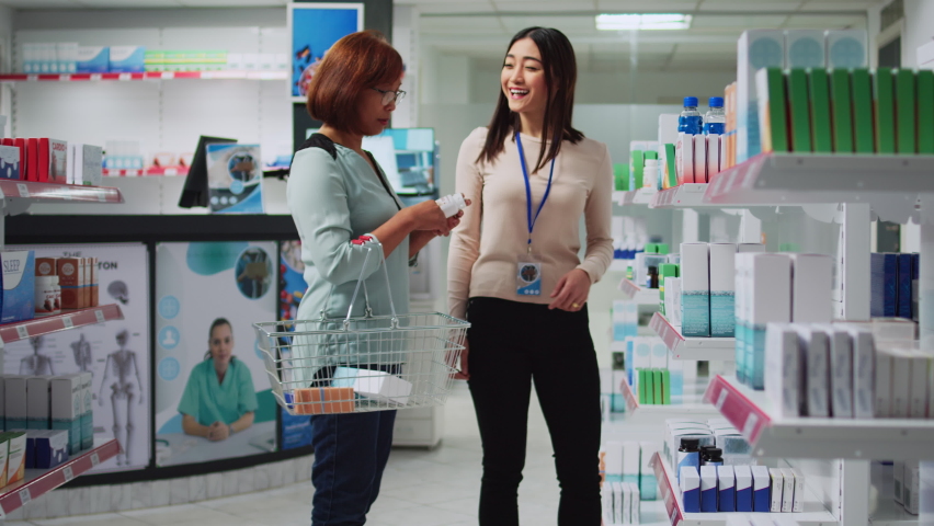 Pharmacy assistant explaining vitamins to female client, talking in pharmaceutical drugstore. Young woman helping customer with medical supplements and health care products, pills packages.