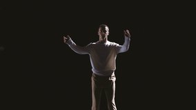 Solo performance of young man in white costume dancing contemp dance in darkness of studio at bright spotlight. Modrn art, action, dance, music, emotions, energy. Video, 4k
