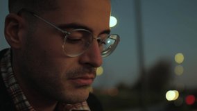 Smooth camera movement of a young and focused man looking at the horizon while flying his drone using a remote controller connected to his smartphone at dusk or sunset. Bokeh lights in the background.