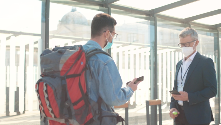 Side view of Caucasian man in mask travelling scanning qr code showing vaccination certificate on smartphone on street. Male measuring temperature of guy tourist with backpack, vaccine passport