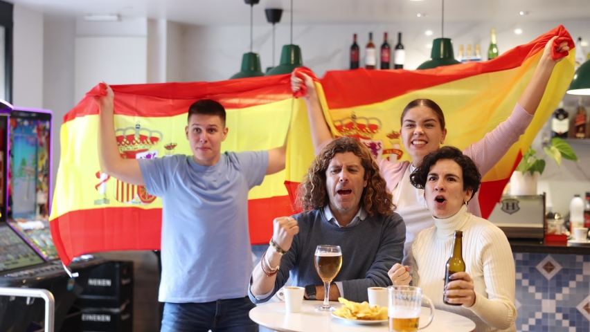 Cheerful footbal fans waving the flag of Spain while drinking beer and watching tournament in sport bar