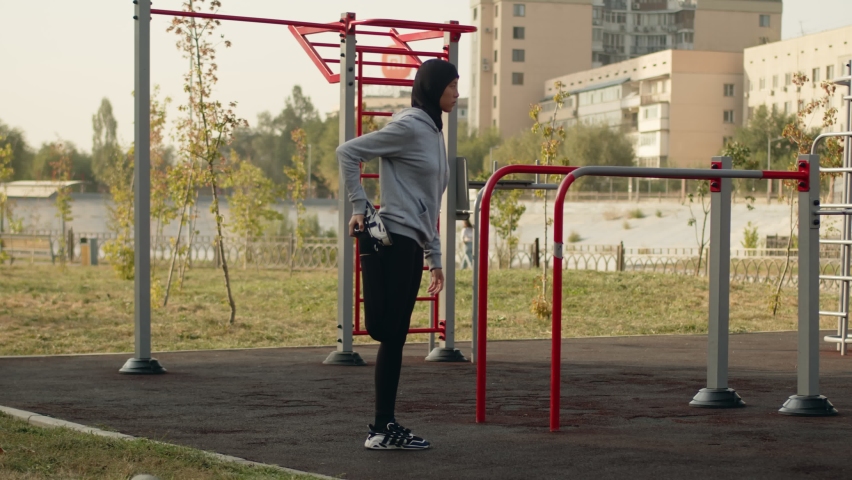 Muslim girl goes in for sports in the city park outdoors. The sportswoman stretches her legs before running, jogging. A woman in a black hijab and a gray sweatshirt on a sports ground. | Shutterstock HD Video #1097500241