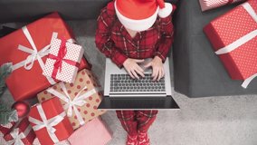 New Year holidays shopping and ordering. A girl in Santa Claus hat communicates via video call on computer. Celebrating home online. Woman in red on sofa at home with laptop, Christmas tree and gifts.