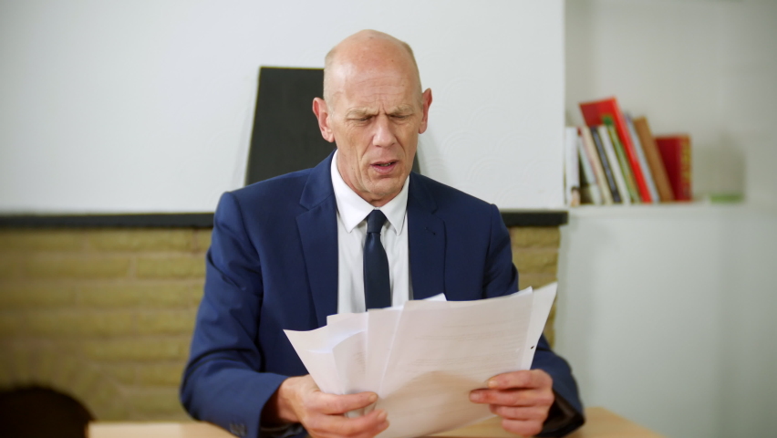 A depressed businessman is sitting at a desk looking at paperwork. The man is stressed and holds his head in his hands. | Shutterstock HD Video #1097504063